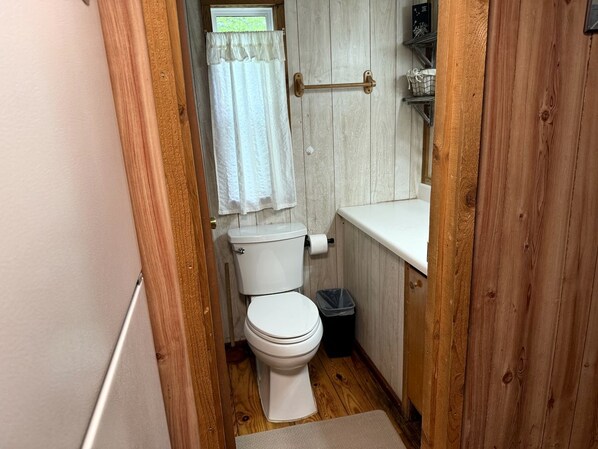 A small bathroom with a toilet, a trash bin, a towel rack, and a window with a curtain.