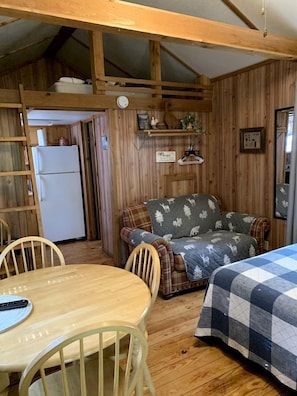 A cozy cabin interior with a wooden dining table, a sofa with a patterned blanket, a bed with a checkered blanket, and a refrigerator in the background.
