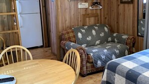 Un interior acogedor de cabaña con una mesa de comedor de madera, un sofá con una manta estampada, una cama con una manta a cuadros y un refrigerador al fondo.