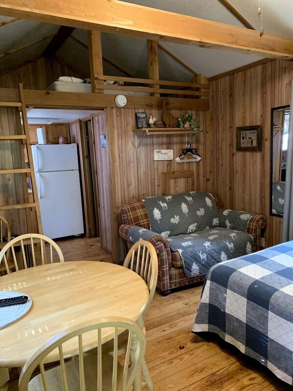 Un interior acogedor de cabaña con una mesa de comedor de madera, un sofá con una manta estampada, una cama con una manta a cuadros y un refrigerador al fondo.