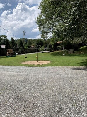 Un espacio cubierto de césped con un campo de béisbol, un conjunto de columpios de madera y un árbol.