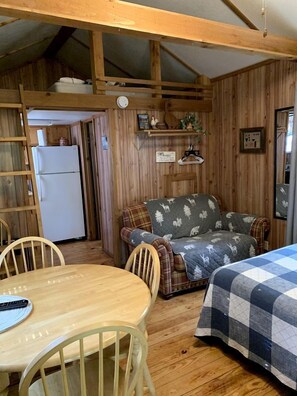 A cozy cabin interior with a wooden dining table, a sofa with a patterned blanket, a bed with a checkered blanket, and a refrigerator in the background.