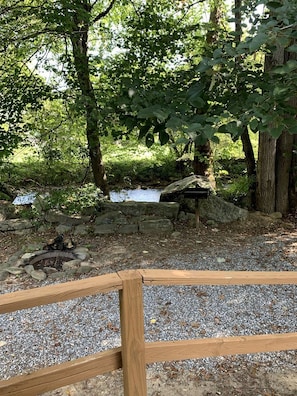 A natural outdoor setting with a wooden fence, a small stream, and a stone fire pit.