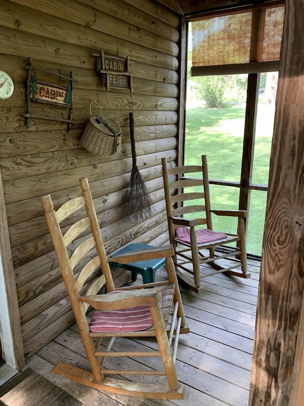 A cabin interior with wooden walls, a rocking chair, a table, and a window with a view of greenery.