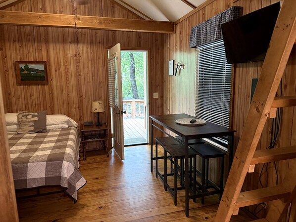 A wooden cabin interior with a bed, a table with chairs, a TV, and a door leading outside.