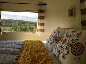 A bedroom with a bed, a window with curtains, and a view of greenery outside. - Charming Riverfront Airstream Rental for Getaway near Fredericksburg, Texas (Mason)