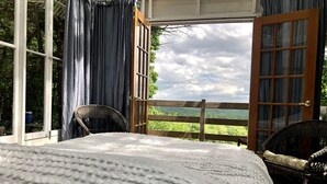 Un dormitorio con una cama grande, una silla de mimbre y vistas a un paisaje verde a través de puertas abiertas.