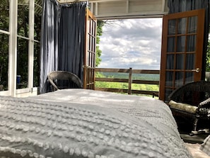 Un dormitorio con una cama grande, una silla de mimbre y vistas a un paisaje verde a través de puertas abiertas.