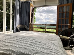 A bedroom with a large bed, a wicker chair, and a view of a green landscape through open doors.