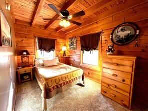 A wooden cabin room with a bed, a dresser, a ceiling fan, and a window with curtains. - Charming Cabin Rental with a Hot Tub and Fireplace in the Smoky Mountains (Clyde)