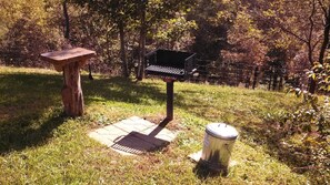 Ein Grill, ein Holztisch und eine Mülltonne in einer grünen Außenanlage.