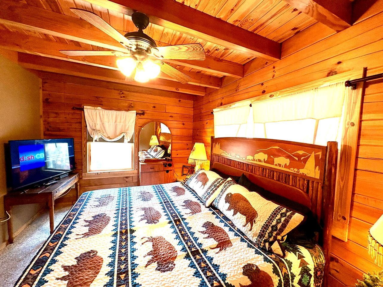 Un dormitorio de cabaña de madera con cama, televisor, ventilador de techo y ventana con cortinas.