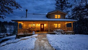 Una cabaña de troncos con un porche, una chimenea y un paisaje cubierto de nieve.