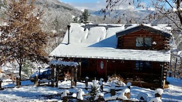 Una baita di legno con tetto innevato, circondata da alberi e con montagne sullo sfondo.