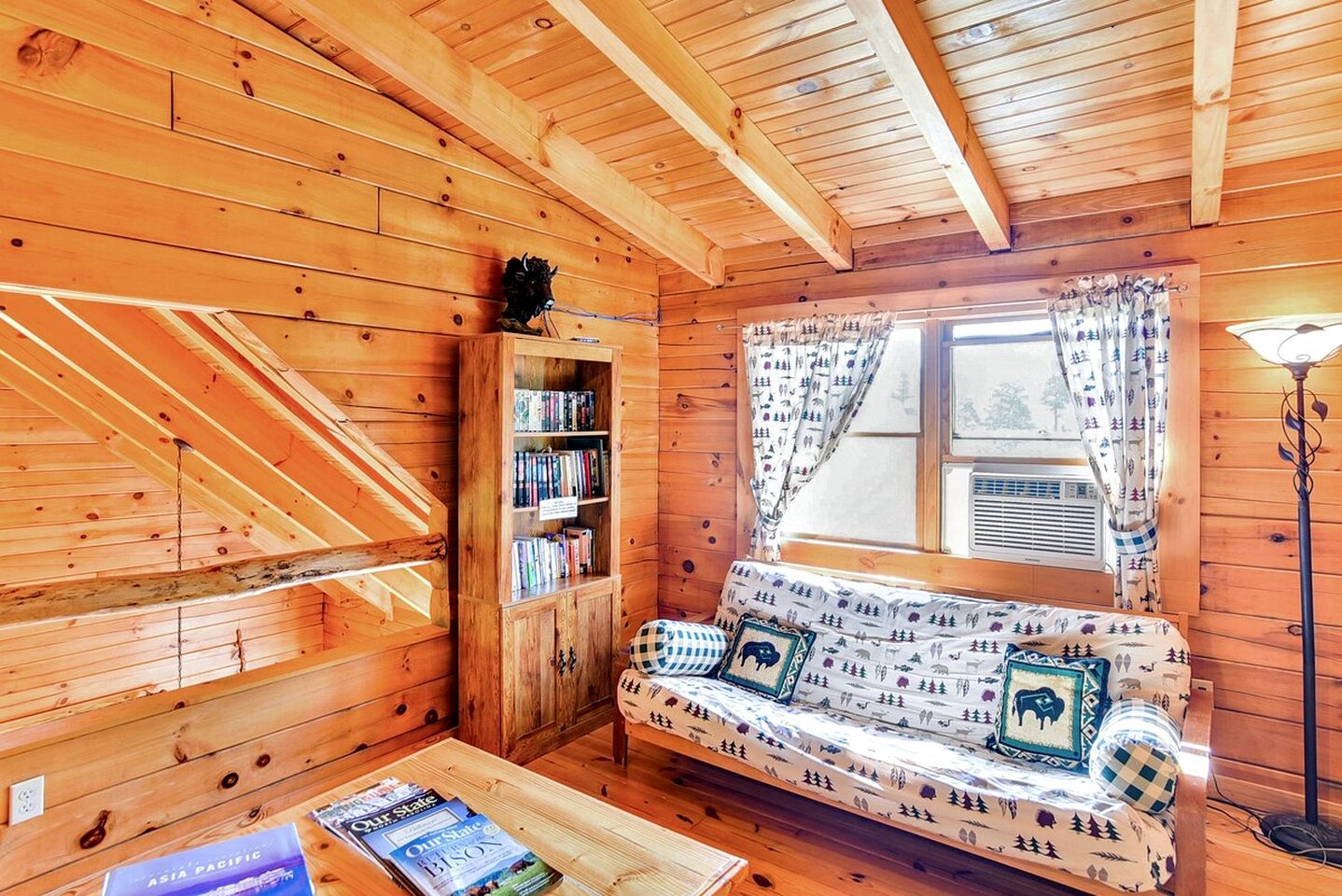 A cozy cabin interior with a wooden staircase, a bookshelf, a sofa with patterned cushions, and a window with curtains.