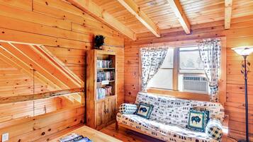 A cozy cabin interior with a wooden staircase, a bookshelf, a sofa with patterned cushions, and a window with curtains.