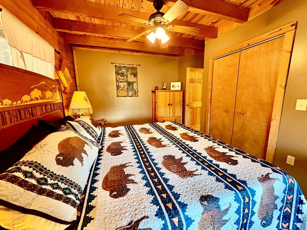 A room with a bed covered in a patterned quilt, wooden walls, a ceiling fan, and a door.