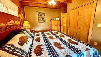 A room with a bed covered in a patterned quilt, wooden walls, a ceiling fan, and a door.