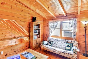 A cozy cabin interior with a wooden staircase, a bookshelf, a sofa with patterned cushions, and a window with curtains.