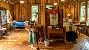 Interior rústico de cabana com teto de madeira, uma cama, uma mesa com abajur e uma janela com vista para vegetação.