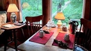 Uma mesa de jantar de madeira com cadeiras, duas lâmpadas e vista para uma floresta através de grandes janelas.