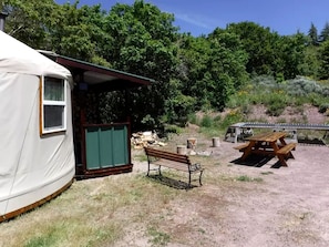 Una cabaña rústica con banca de madera y mesa de picnic, rodeada de pastizal seco y árboles.