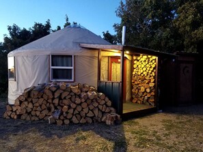 Une yourte, avec une pile de bois de chauffage à l’extérieur, une fenêtre et une porte donnant sur une pièce dont un mur est recouvert de bûches empilées.