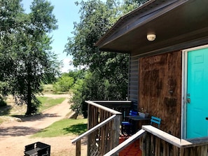 A wooden porch with a blue door, a small table with chairs, and a pathway leading to a dirt road. - Bright and Cheery Riverside Cabin in Texas Wine Country (Mason)