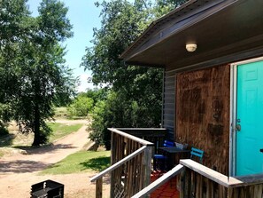 A wooden porch with a blue door, a small table with chairs, and a pathway leading to a dirt road. - Bright and Cheery Riverside Cabin in Texas Wine Country (Mason)