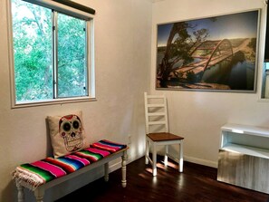 Una habitación con un banco, una silla, un cuadro y una ventana con vista a árboles.