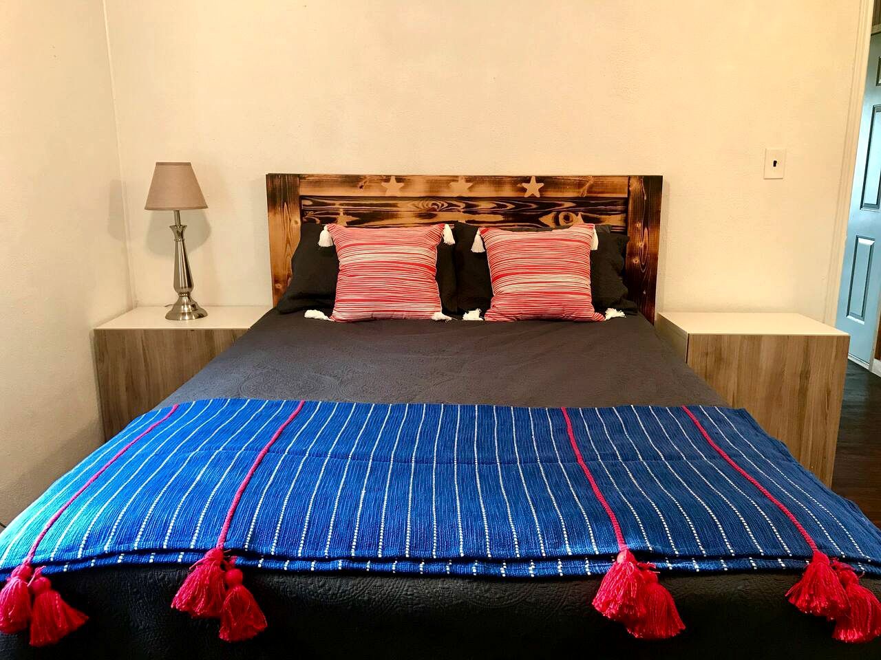Een bed met een blauw gestreept laken en rode kwastjes, twee kussens, een houten hoofdbord en een bedlampje.