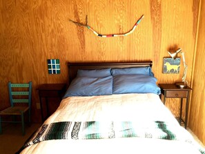 Una habitación con paneles de madera, una cama, una silla, una mesita y un adorno en la pared.
