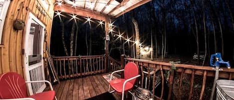 Una terraza de madera con barandilla, dos sillas rojas, un calentador metálico y vista a un bosque de noche.