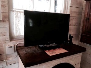 Une pièce avec un bureau en bois, un téléviseur noir, un décodeur Samsung et une fenêtre ornée de rideaux à motifs.