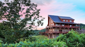 A large wooden cabin with multiple balconies, surrounded by lush greenery and a scenic view.