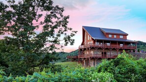 A large wooden cabin with multiple balconies, surrounded by lush greenery and a scenic view.
