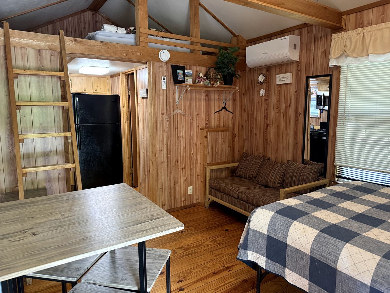 A compact room with a bunk bed, a small table, a refrigerator, and a window with blinds.