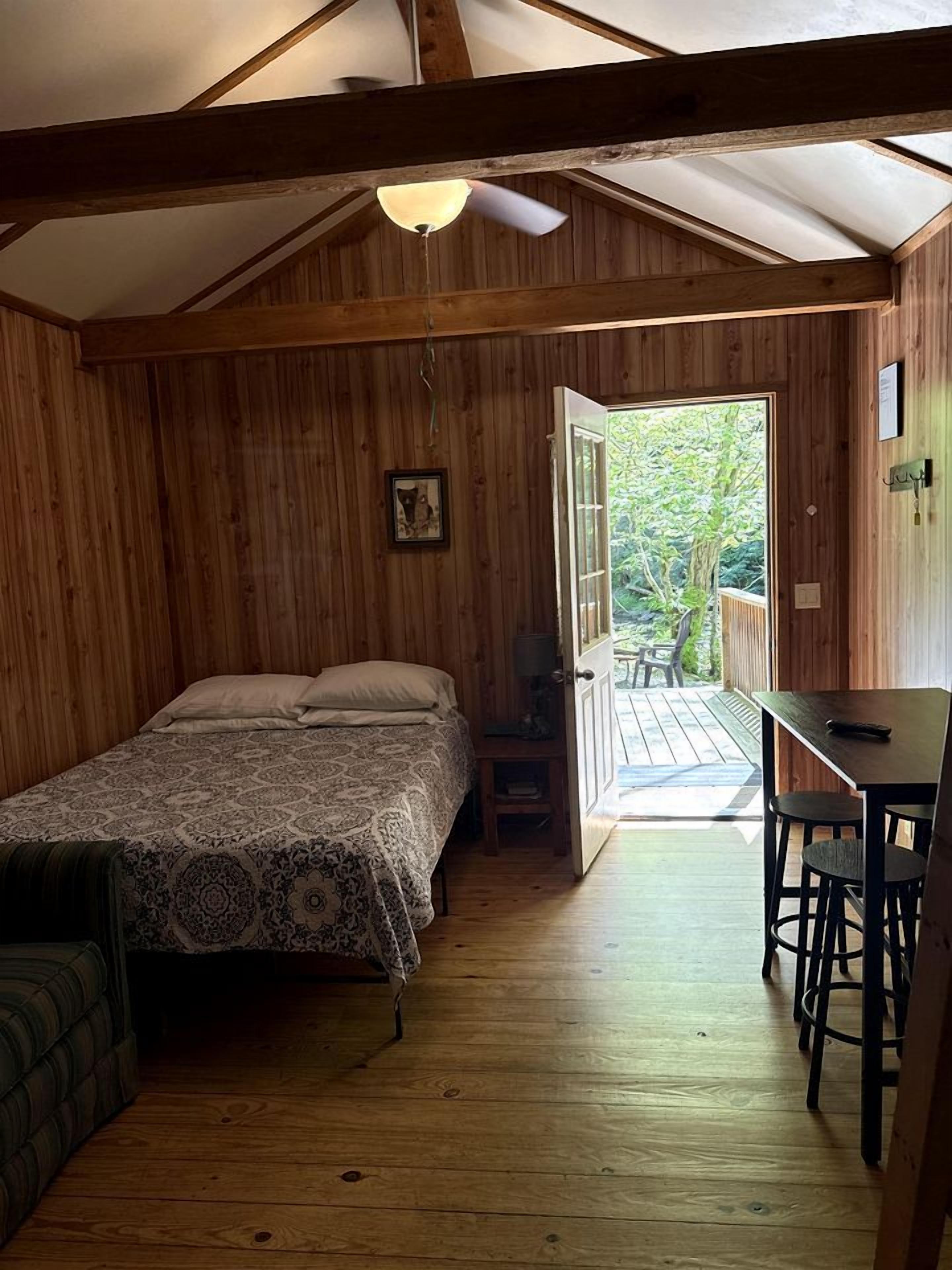 A wooden room with a bed, a table, and a chair. There is a door leading outside.