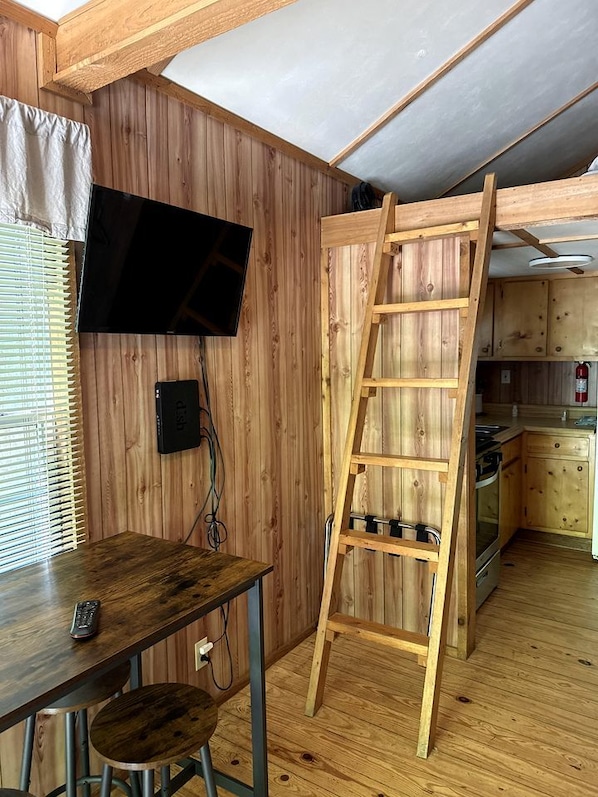Une pièce avec un intérieur en bois, un téléviseur fixé au mur, une échelle en bois, un coin cuisine avec des placards et une cuisinière, ainsi qu’une table avec une chaise.