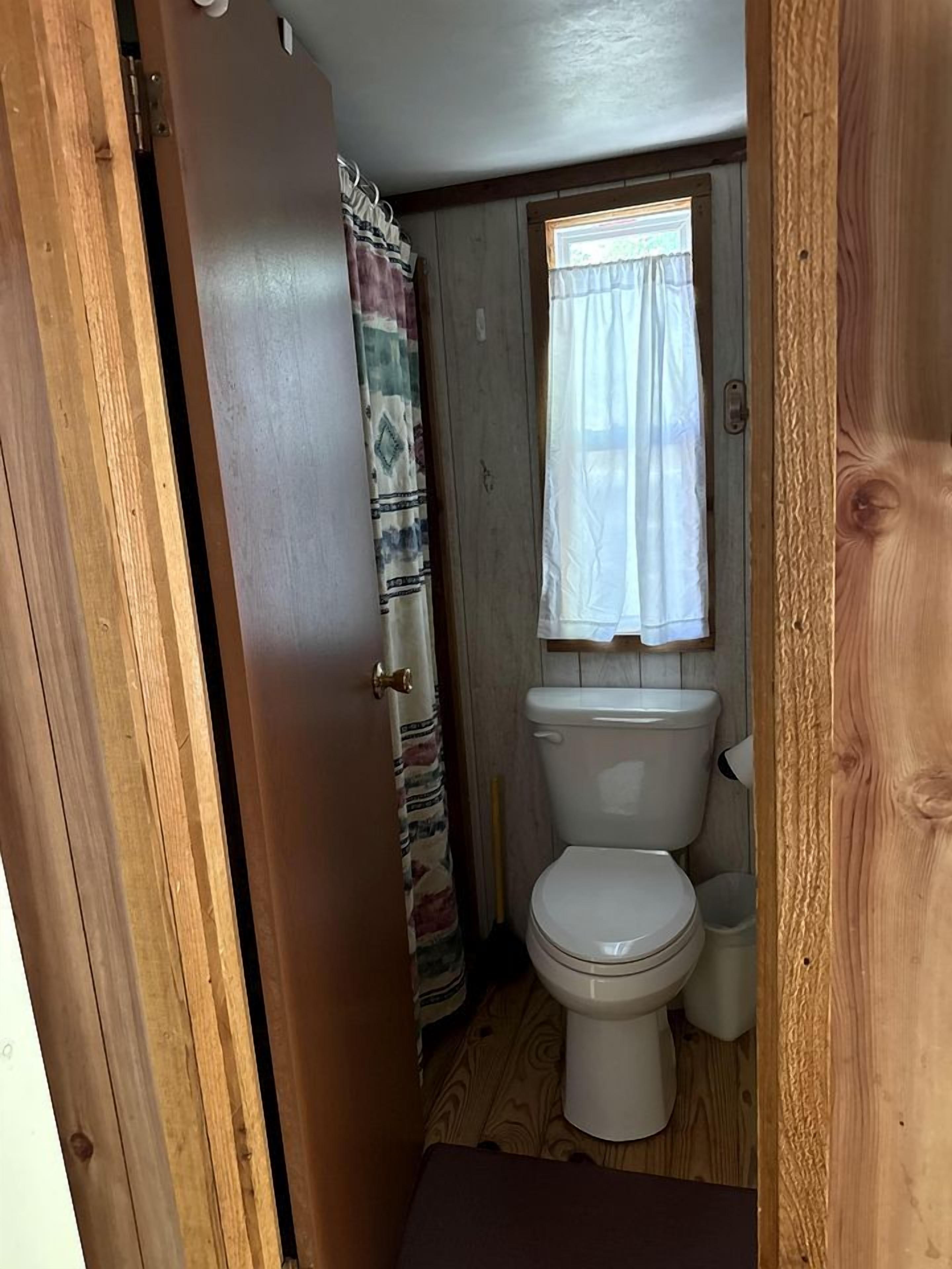 Eine Holztür führt zu einem Badezimmer mit Toilette, einem Fenster mit Vorhang und einem Holzboden.