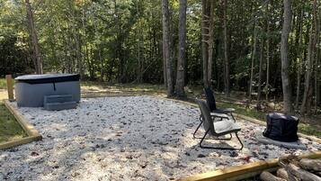 A gravel patio with a chair, a fire pit, and a trash can in a wooded area.