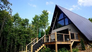 A cabin with a steeply pitched roof, large windows, and a wooden deck with stairs leading to the entrance.