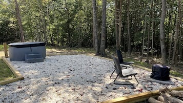 A gravel patio with a chair, a fire pit, and a trash can in a wooded area.