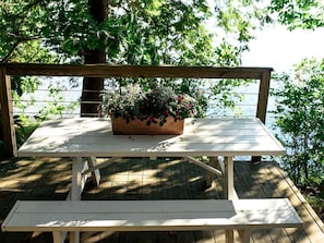 A wooden picnic table with a bench, surrounded by greenery and a wooden deck.