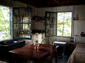 A rustic cabin interior with a wooden table, a chair, a window with a view of trees, and a shelf with various items.