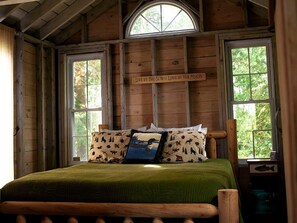A wooden cabin bedroom with a bed, two windows, a wooden ceiling, and a sign above the bed. - Little Diamond Island - on Private Island (West Gardiner)