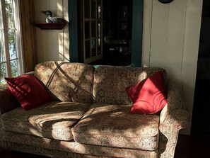 A patterned sofa with two red cushions, a wooden shelf with a decorative item, and a window with sunlight.