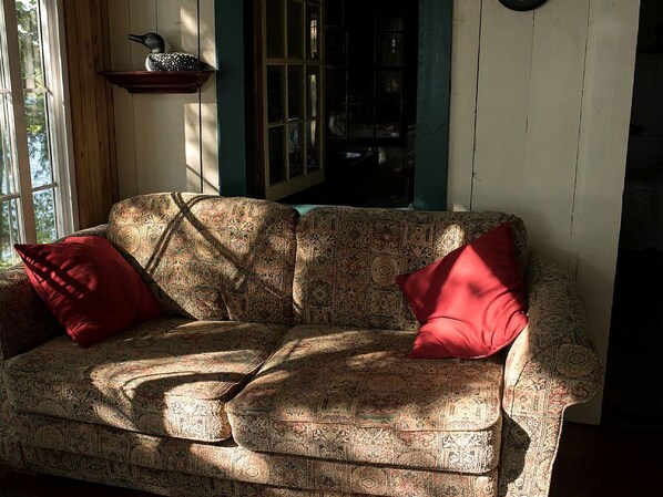 A patterned sofa with two red cushions, a wooden shelf with a decorative item, and a window with sunlight.