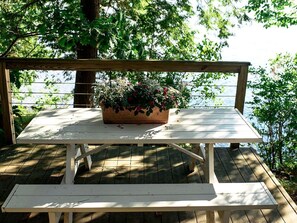 A wooden picnic table with a bench, surrounded by greenery and a wooden deck.