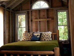 A wooden cabin bedroom with a bed, two windows, a wooden ceiling, and a sign above the bed.